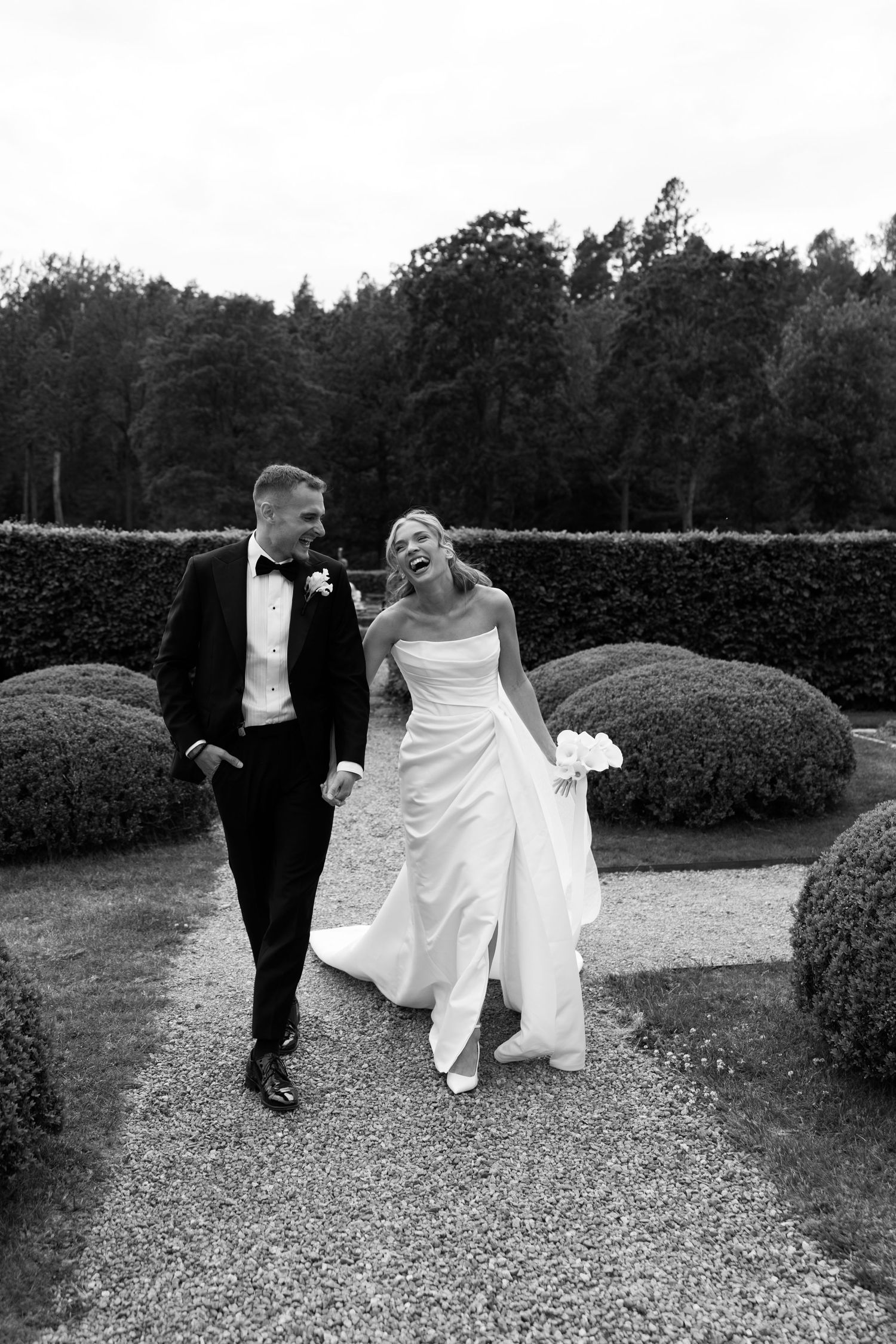 A couple walks together on a gravel path in a formal garden with manicured hedges, captured in black and white.