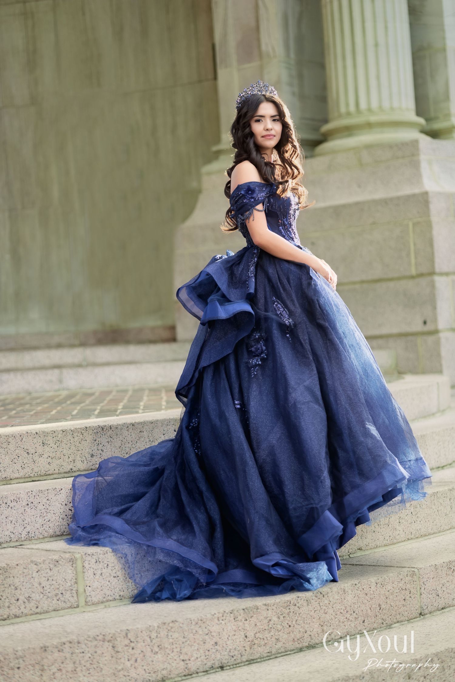 A young girl in a flowing navy blue ball gown poses gracefully on stone steps in an elegant outdoor setting.