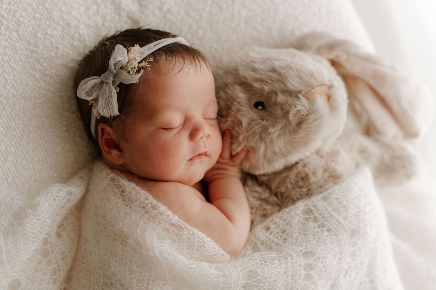 A sleeping newborn baby girl with a floral headband snuggles beside a soft plush bunny.