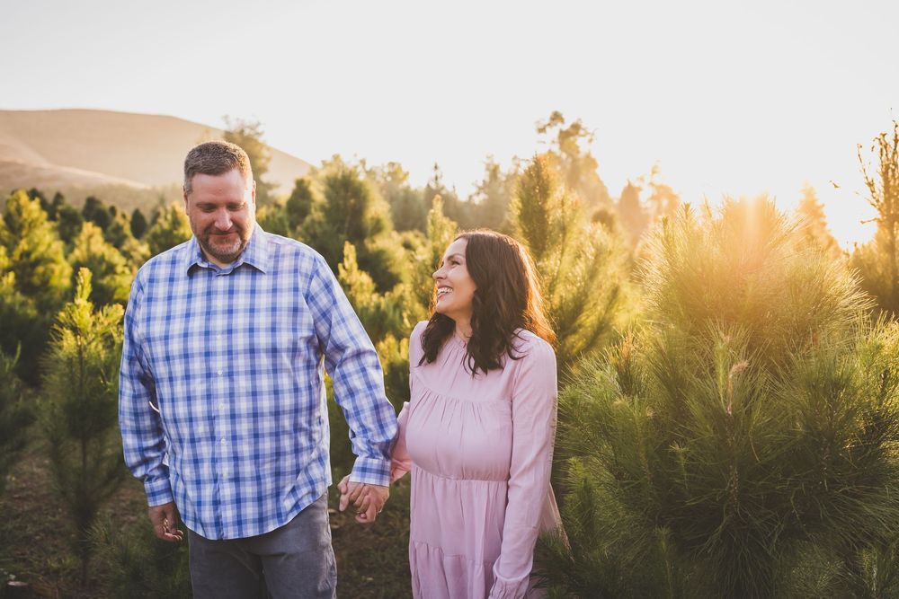 A couple in plaid and pink attire share a romantic moment during golden hour in a pine forest setting.