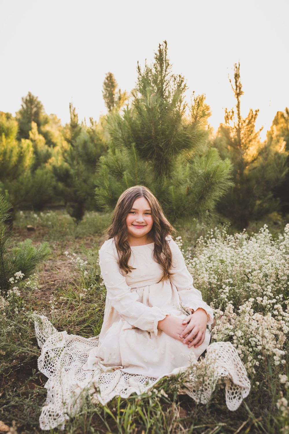 A child in a white lace dress sits among wildflowers and pine trees at golden hour.