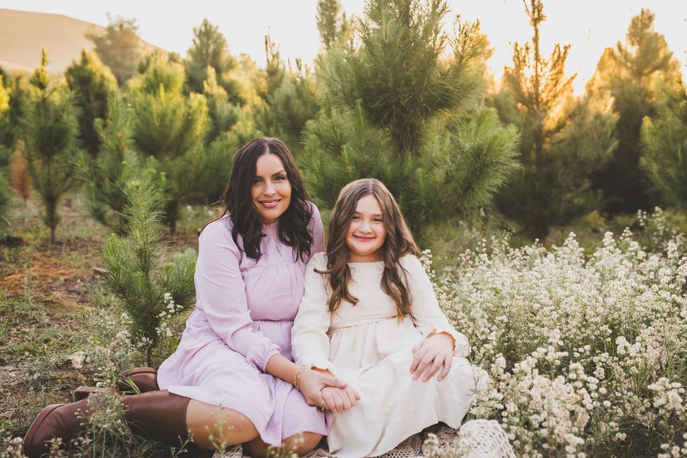 Two people in light colored dresses sit together outdoors among evergreen trees at sunset.