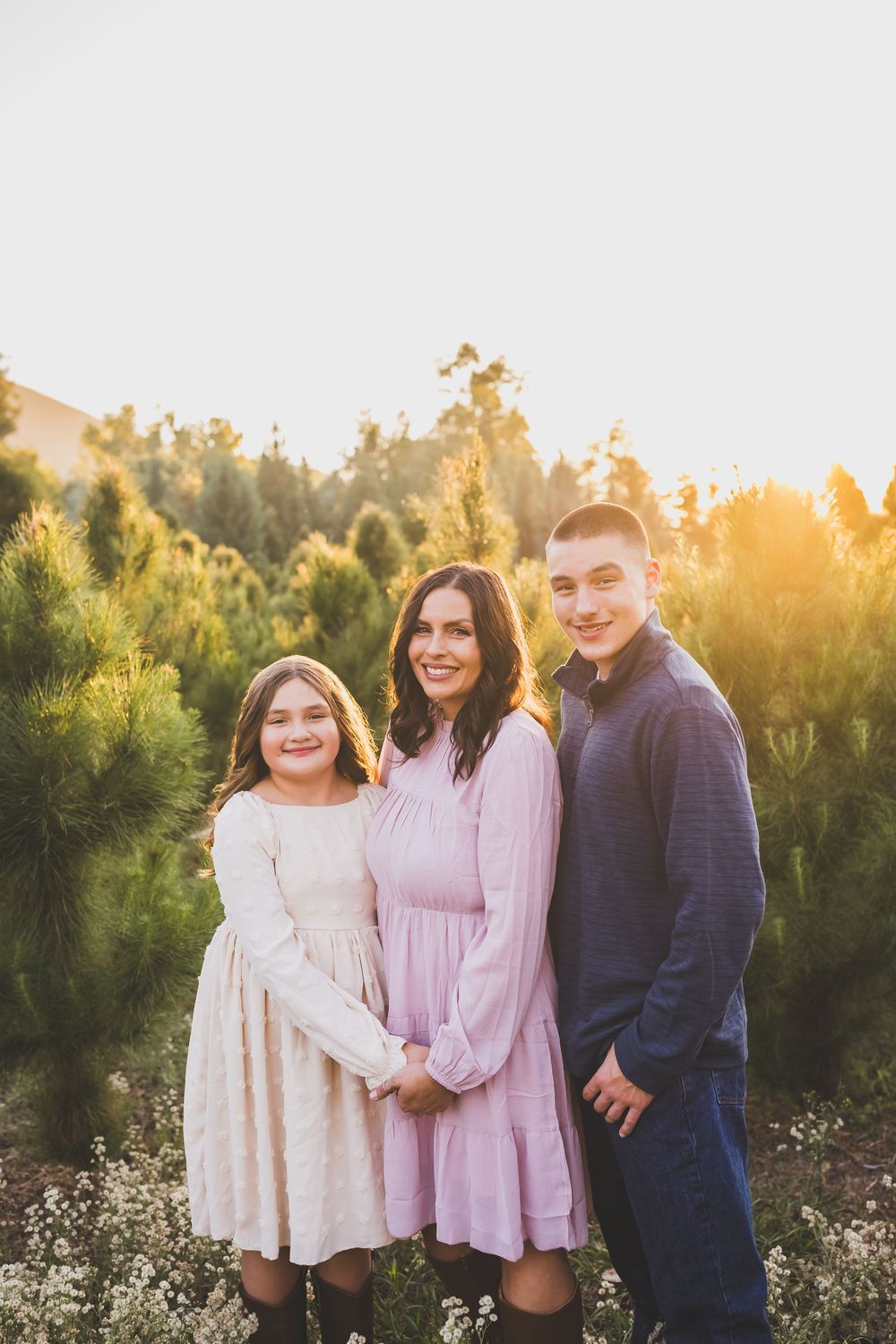 A family poses together in a field at sunset wearing pink and navy outfits among wildflowers and trees.