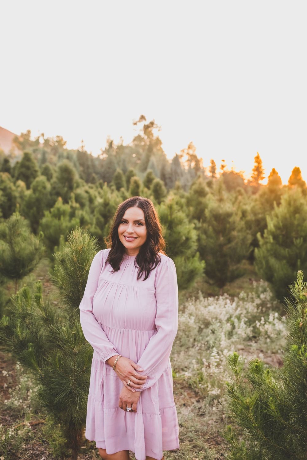 A woman in a flowing pink dress stands in a field of wildflowers and evergreen trees at sunset.