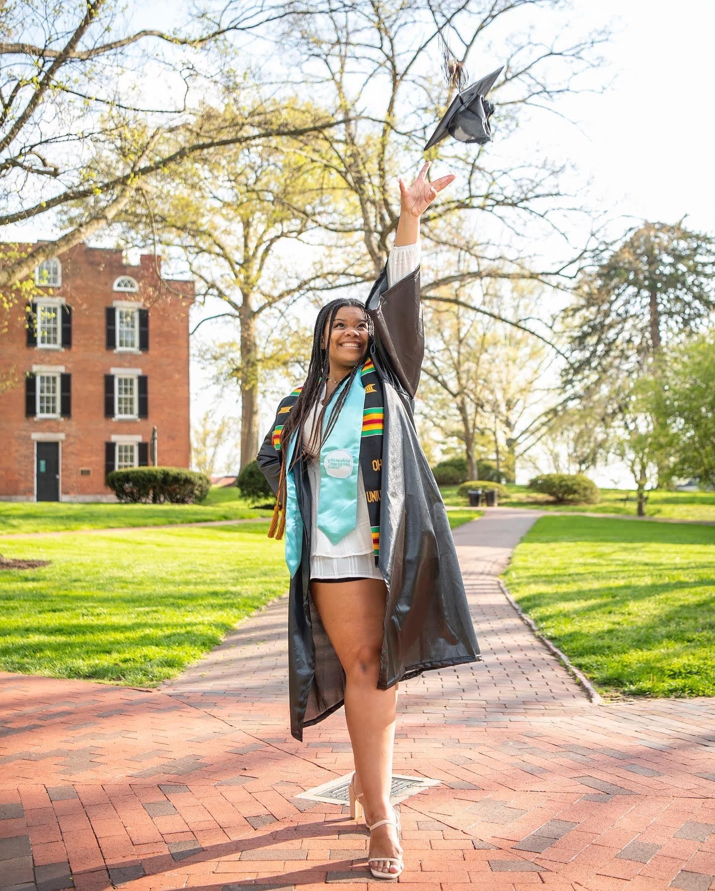 Someone wearing a gray jacket and turquoise top walks along a brick pathway on a sunny spring day near brick buildings.