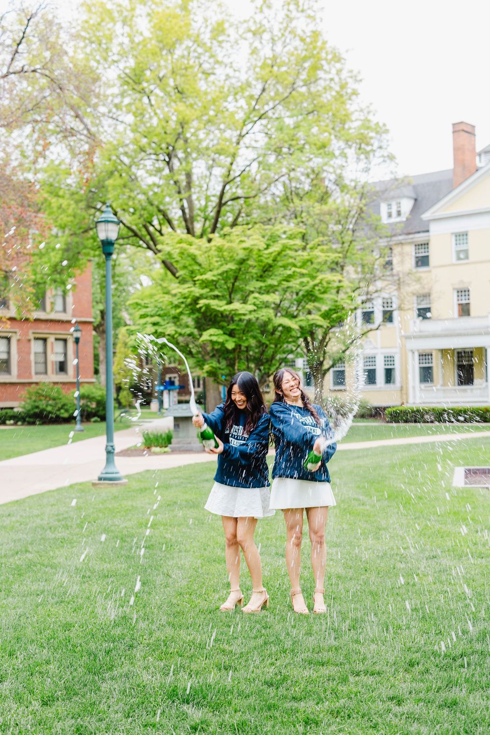 Two young women in navy jackets and white skirts celebrate with confetti on a green campus lawn.
