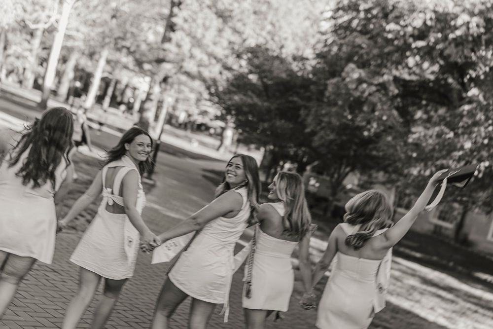 Black and white photo of a group of young women in white dresses laughing and dancing together outdoors.