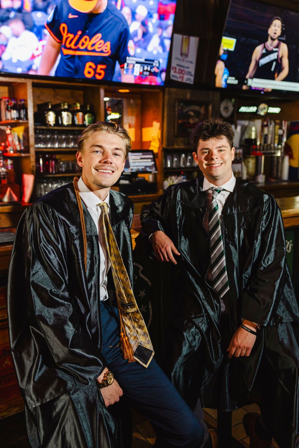 Two smiling graduates in black robes and ties pose together inside a bar with sports TVs in the background.