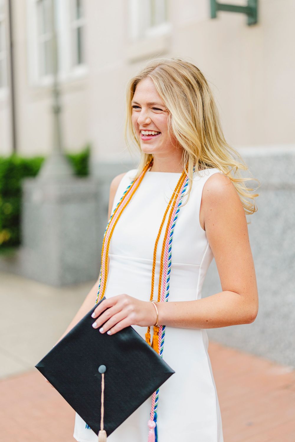 Smiling blonde graduate in white dress holding black cap with colorful honor cords outdoors.