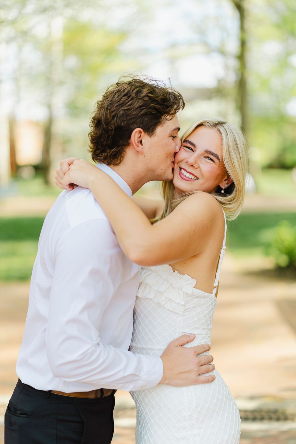 A smiling blonde woman in a white dress receives a kiss on the cheek from a man in a white shirt outdoors.