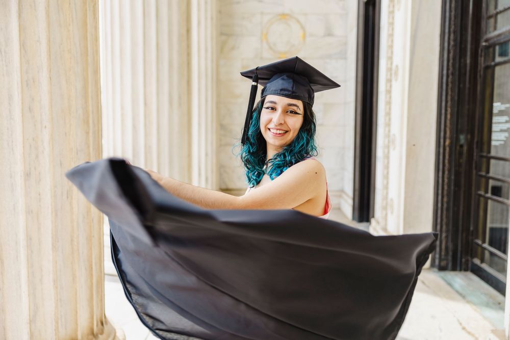Young woman with teal hair in graduation cap and gown smiling outdoors near marble columns.