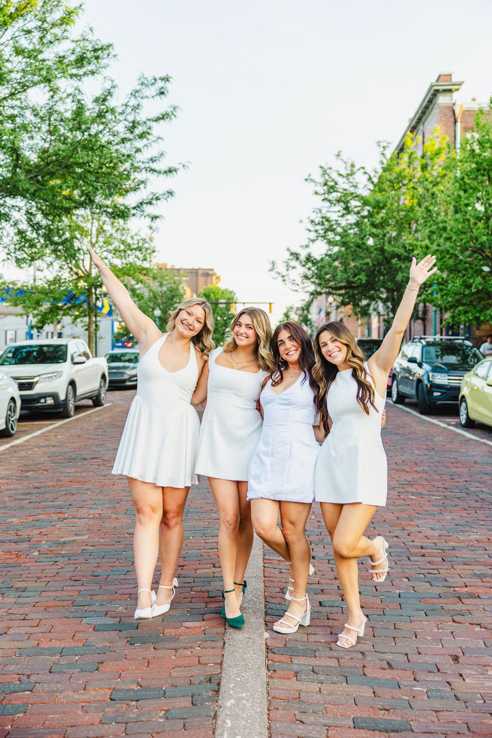 Four women in white dresses pose joyfully on a brick street lined with trees and parked cars.