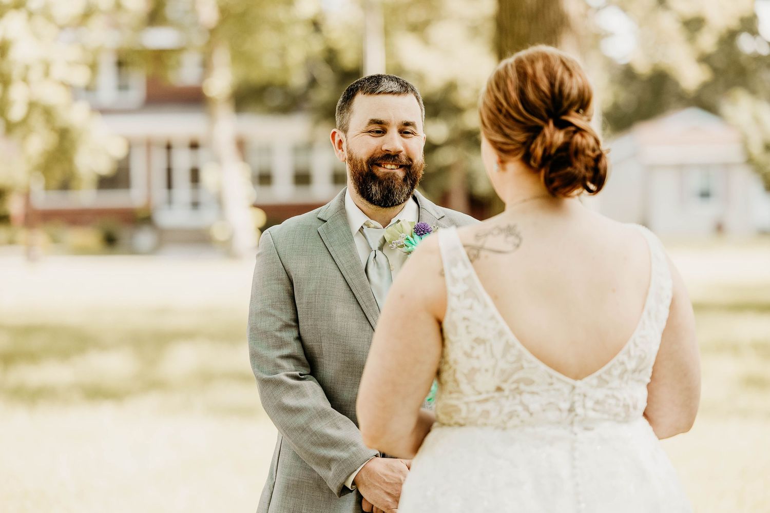 A couple shares a tender moment on their wedding day, with the bride in a lace dress and groom in a grey suit outdoors.