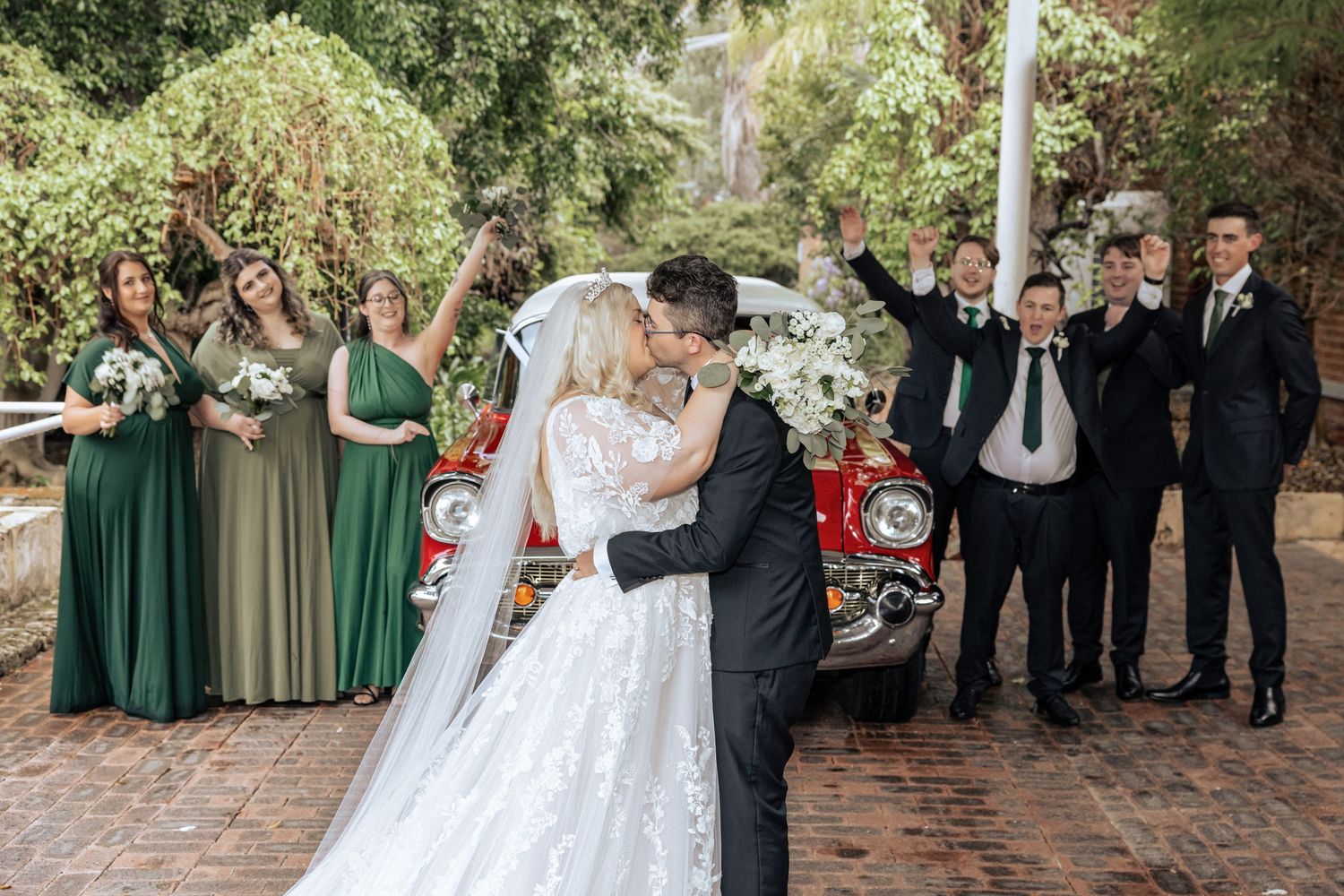 Wedding party members in emerald green dresses and dark suits pose together outdoors for formal photos.