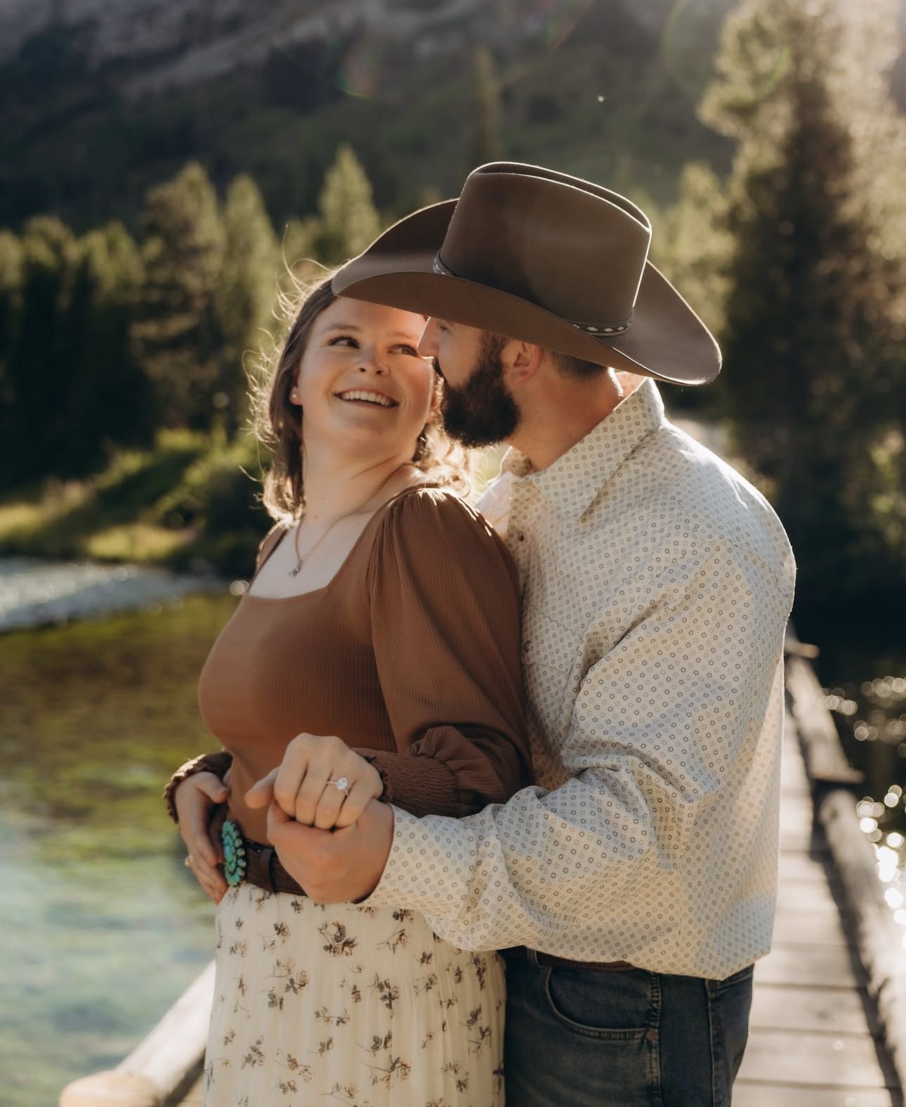A couple in western attire sharing intimate moments on a wooden bridge in a scenic mountain setting.