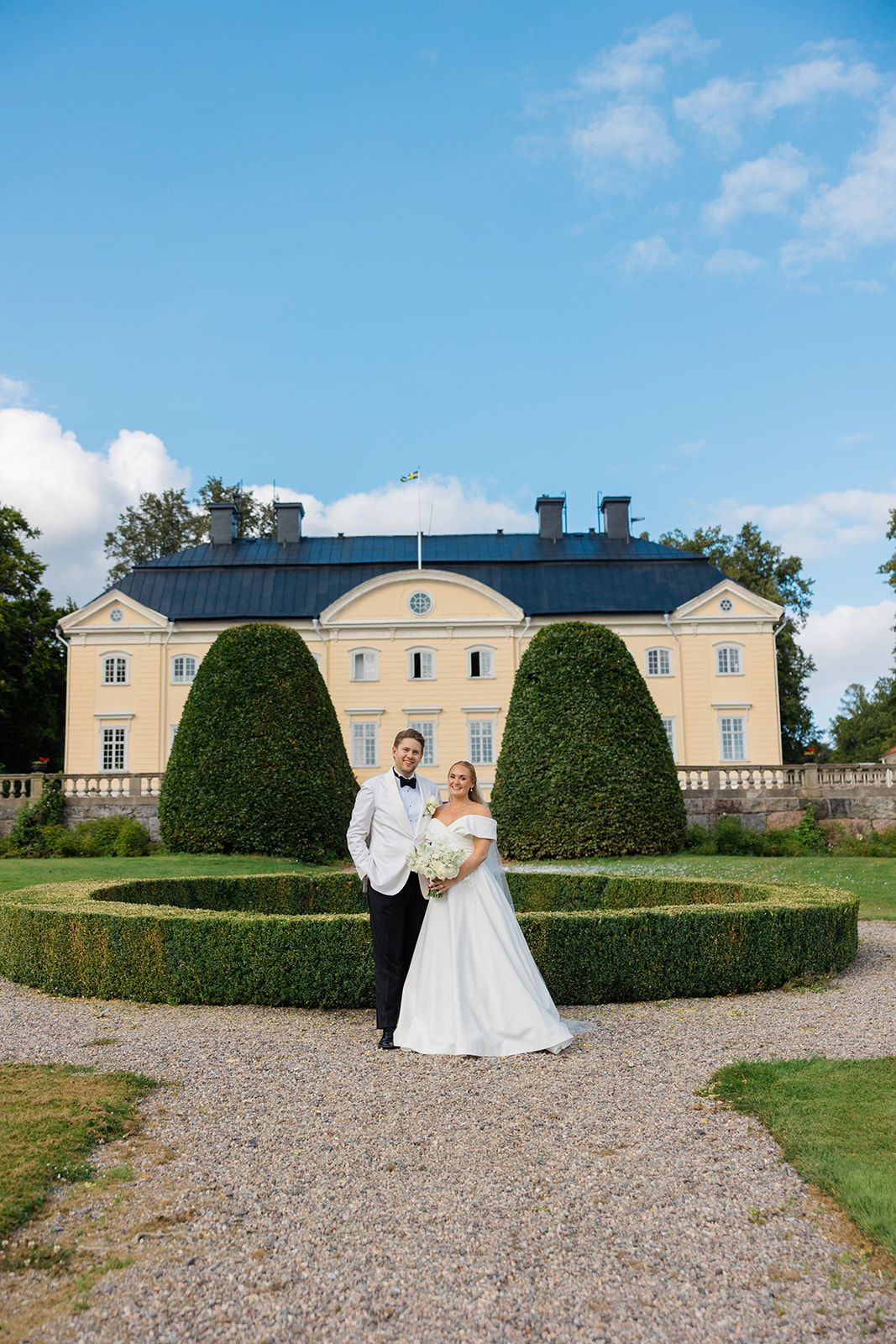 A couple in wedding attire stands on a gravel path in front of a grand yellow mansion with manicured hedges.