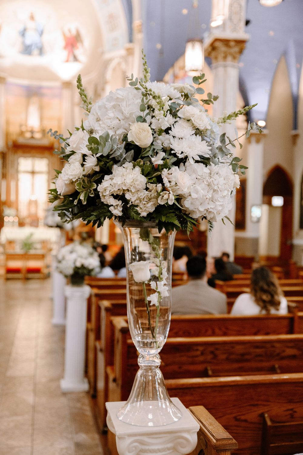 Church aisle decorated with elegant white floral arrangements in glass vases for wedding ceremony.