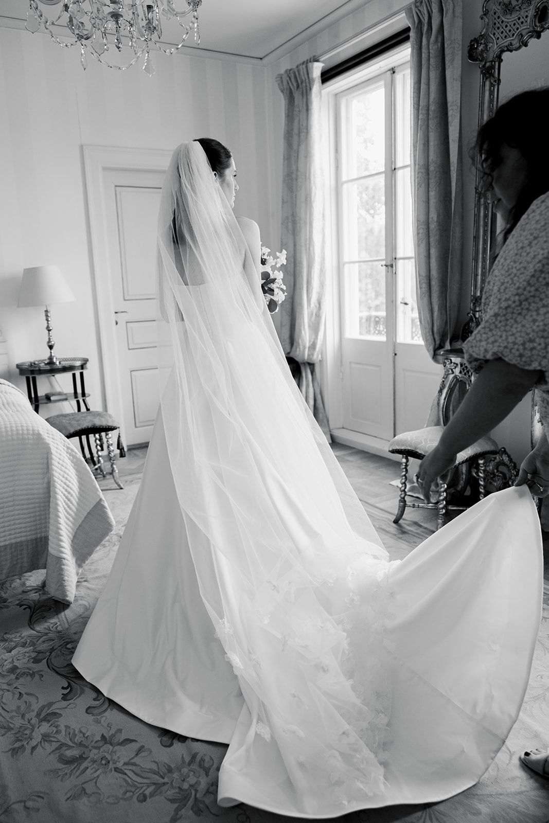 A bride in an elegant white wedding gown with a flowing veil stands in a sunlit room with a crystal chandelier.