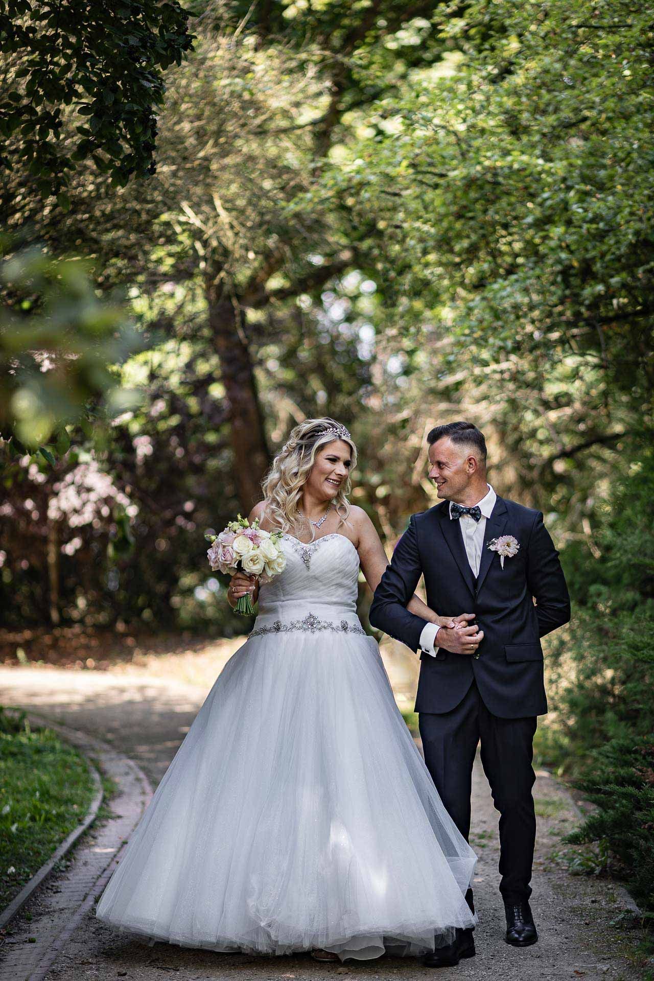 Bride in flowing white gown walks garden path with groom in classic black tuxedo.