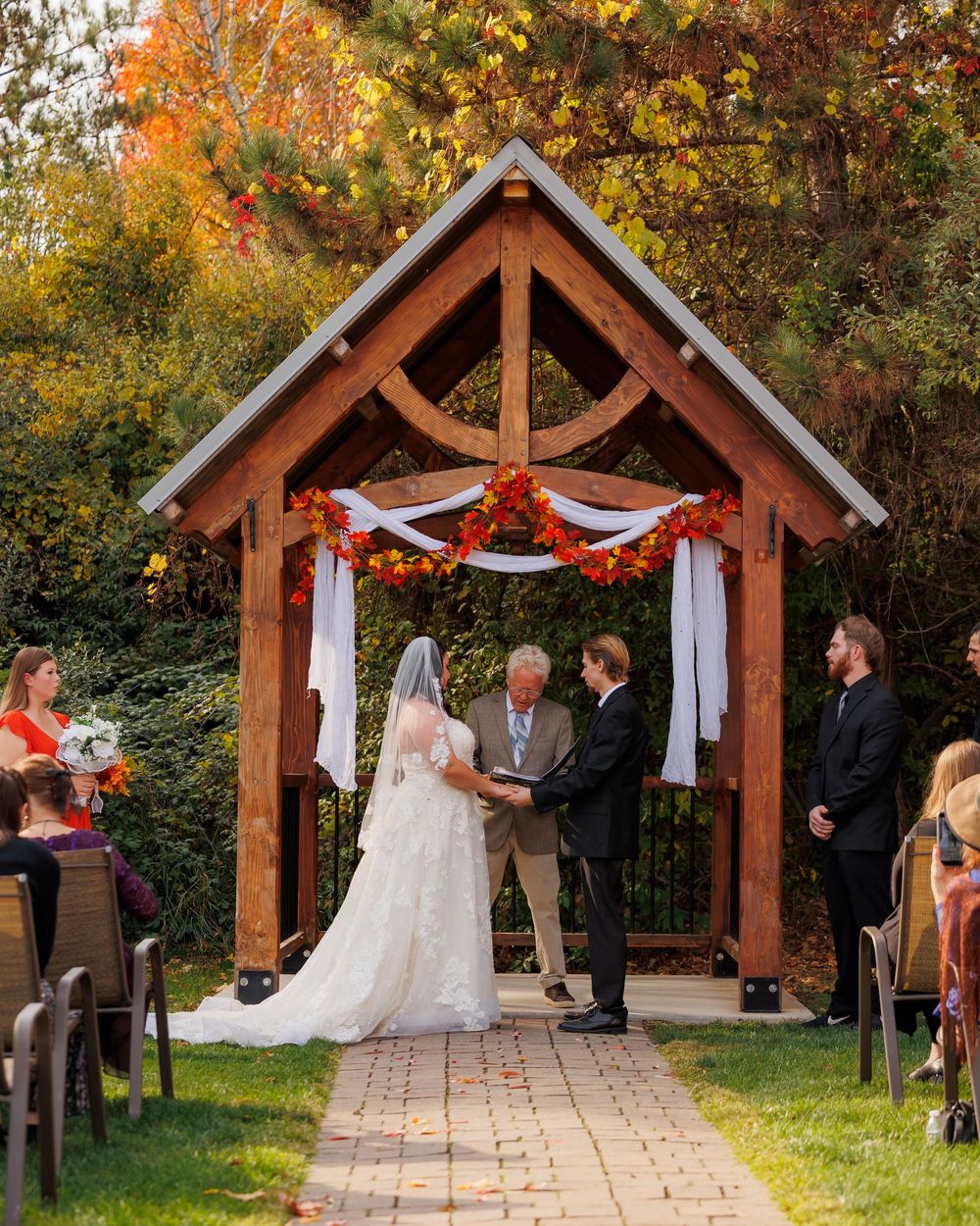 Rustic wooden wedding ceremony arch decorated with white fabric and fall foliage in an outdoor setting.