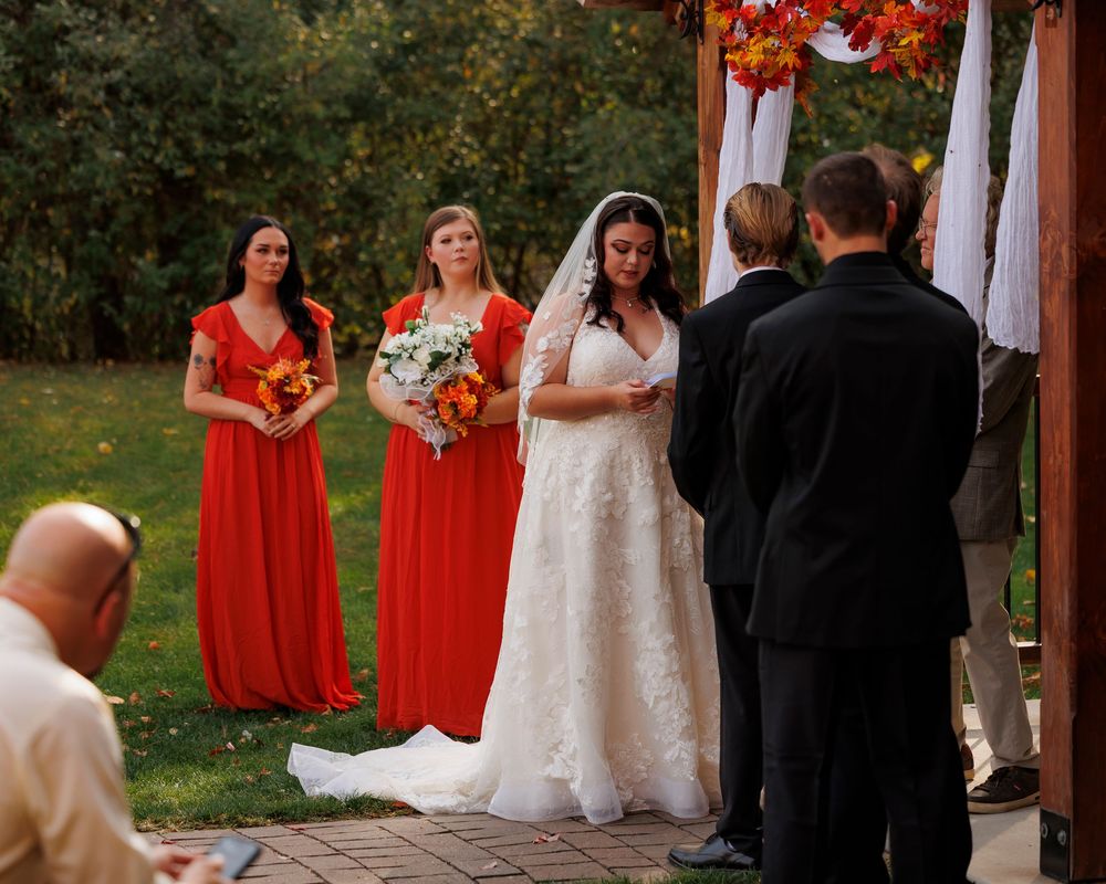 Wedding party members in red dresses and formal attire stand together during an outdoor autumn ceremony.