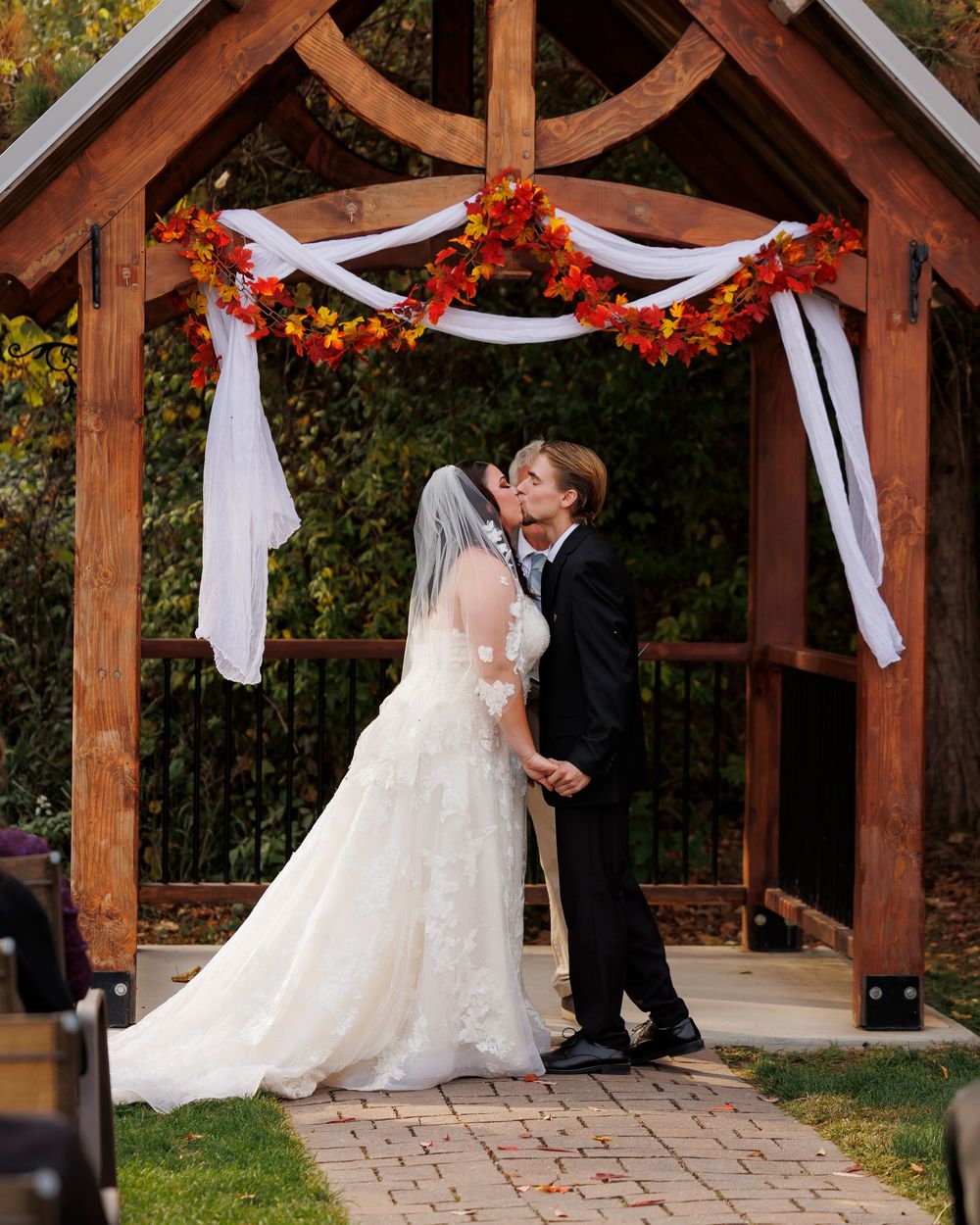 A romantic wedding ceremony takes place under a wooden pergola decorated with autumn flowers and white drapes.