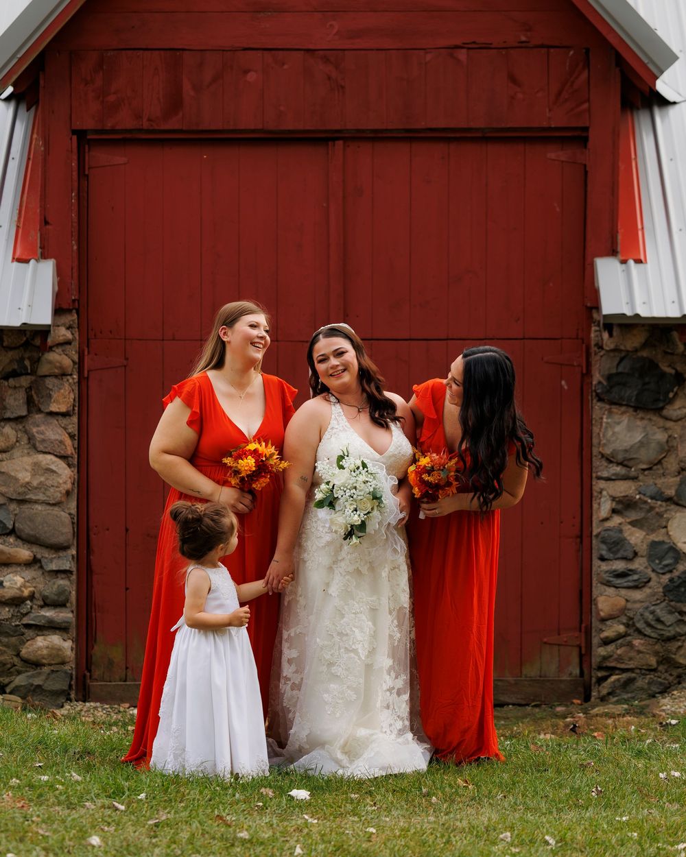 A joyful wedding party poses in front of a rustic red barn with stone foundation and autumn flowers.