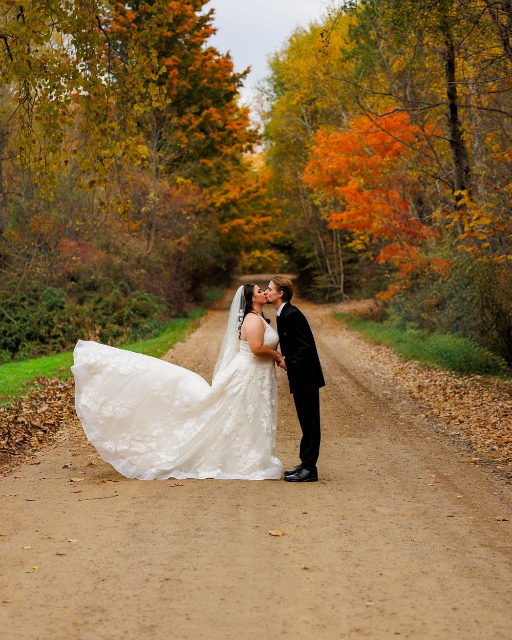 A wedding couple poses on a dirt path surrounded by vibrant autumn foliage.