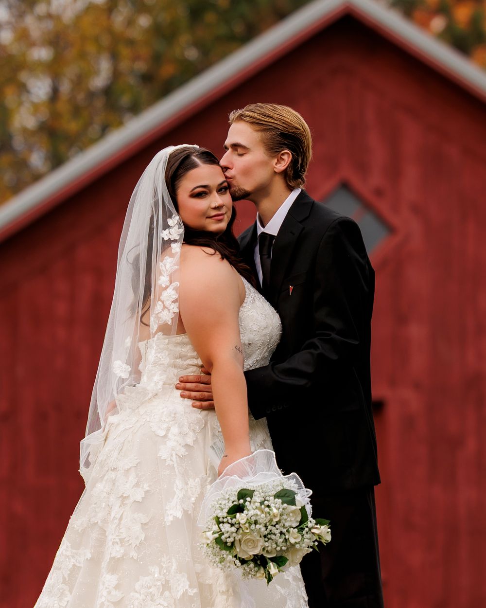 Romantic wedding portraits against a red barn backdrop with fall colors in the background.