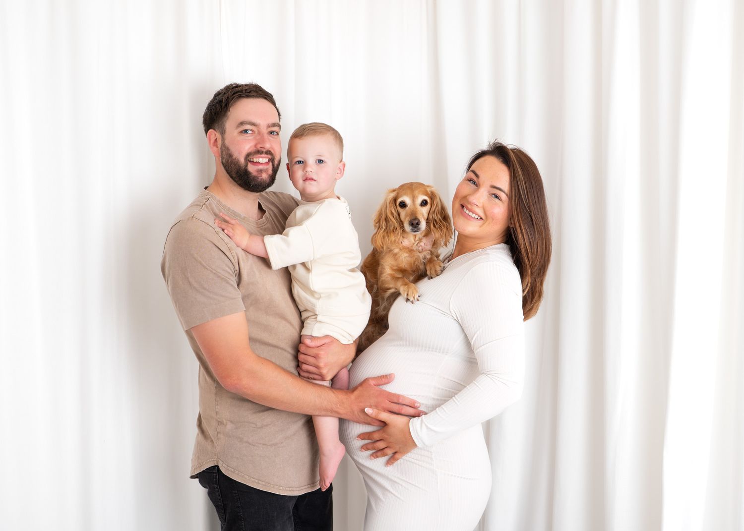 Family in white and beige outfits poses together against a bright white curtain background.