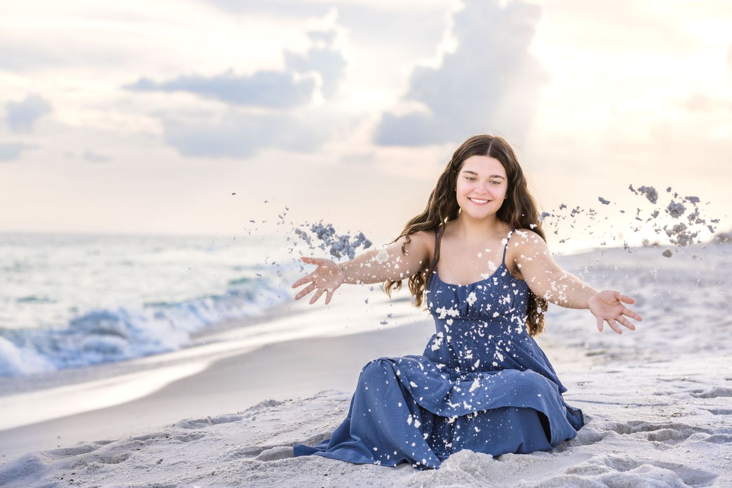Woman in blue dress poses gracefully on white sandy beach with sea grass during golden hour sunset.