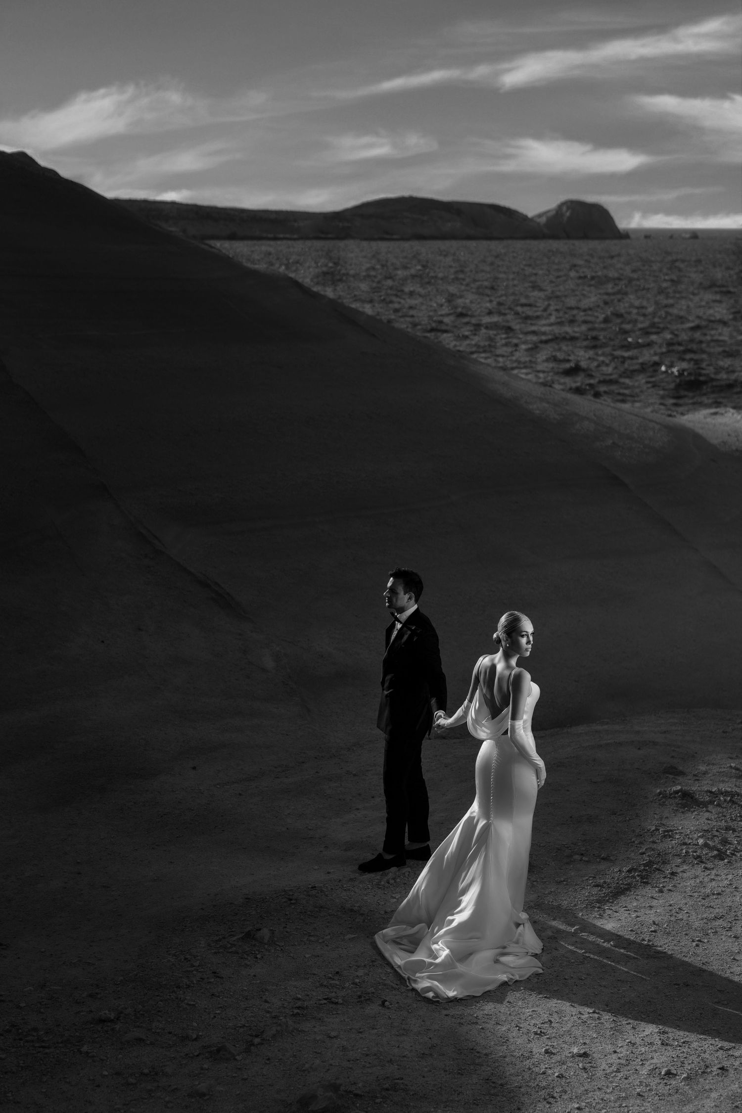 Dramatic black and white wedding photo with couple walking across desert landscape.