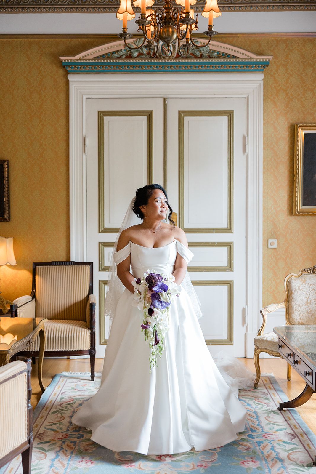 A bride in a white wedding gown poses elegantly in a luxurious hotel room with ornate decor and chandelier lighting.