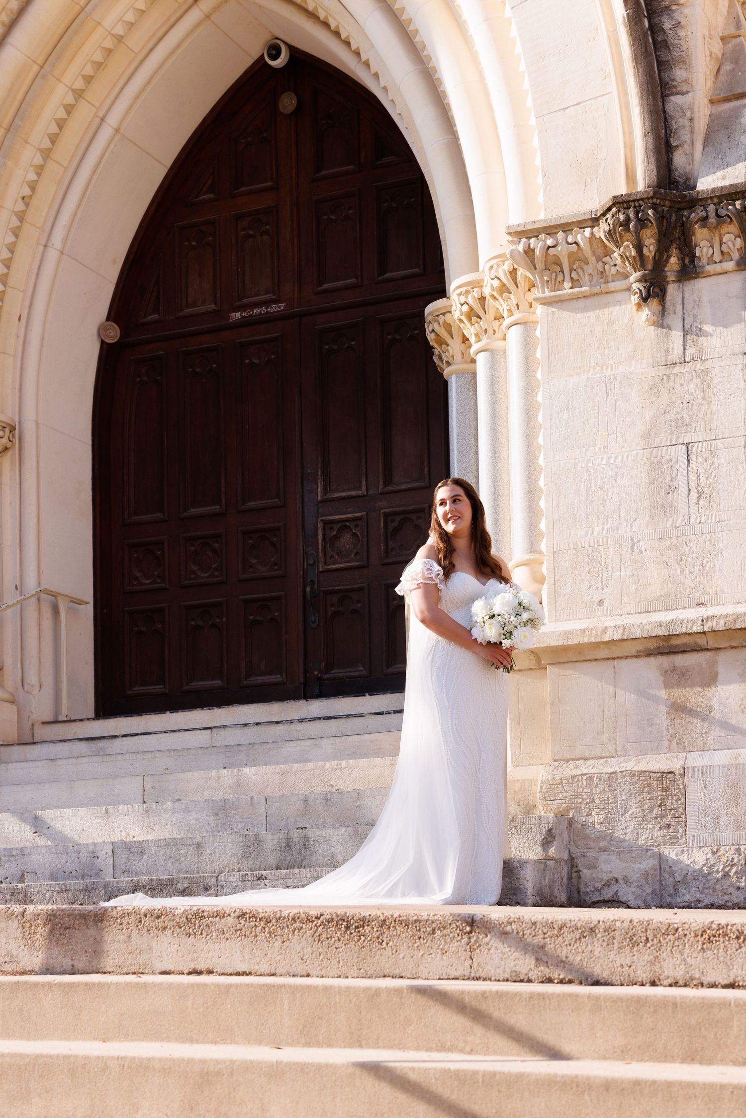 A bride in a white dress holds a bouquet while standing on stone steps in front of an arched church door.