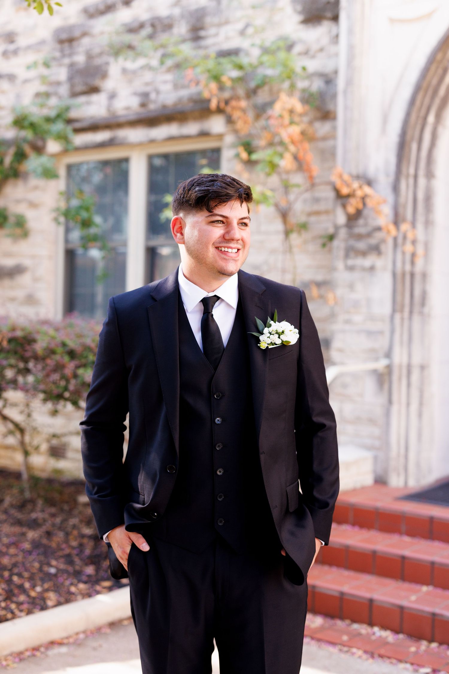 A groom in a classic black tuxedo poses outside a historic stone church building.