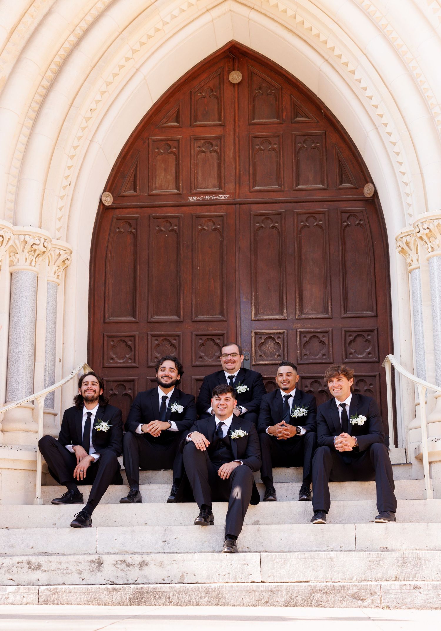 Groomsmen in black suits sit casually on church steps in front of a large wooden door.