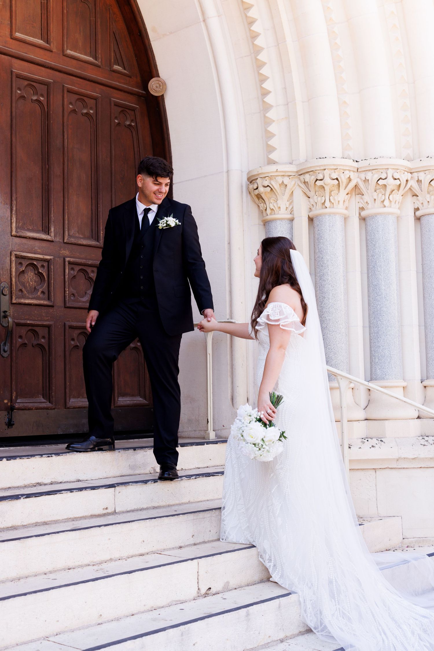 A couple in wedding attire poses on stone steps in front of an ornate church entrance.