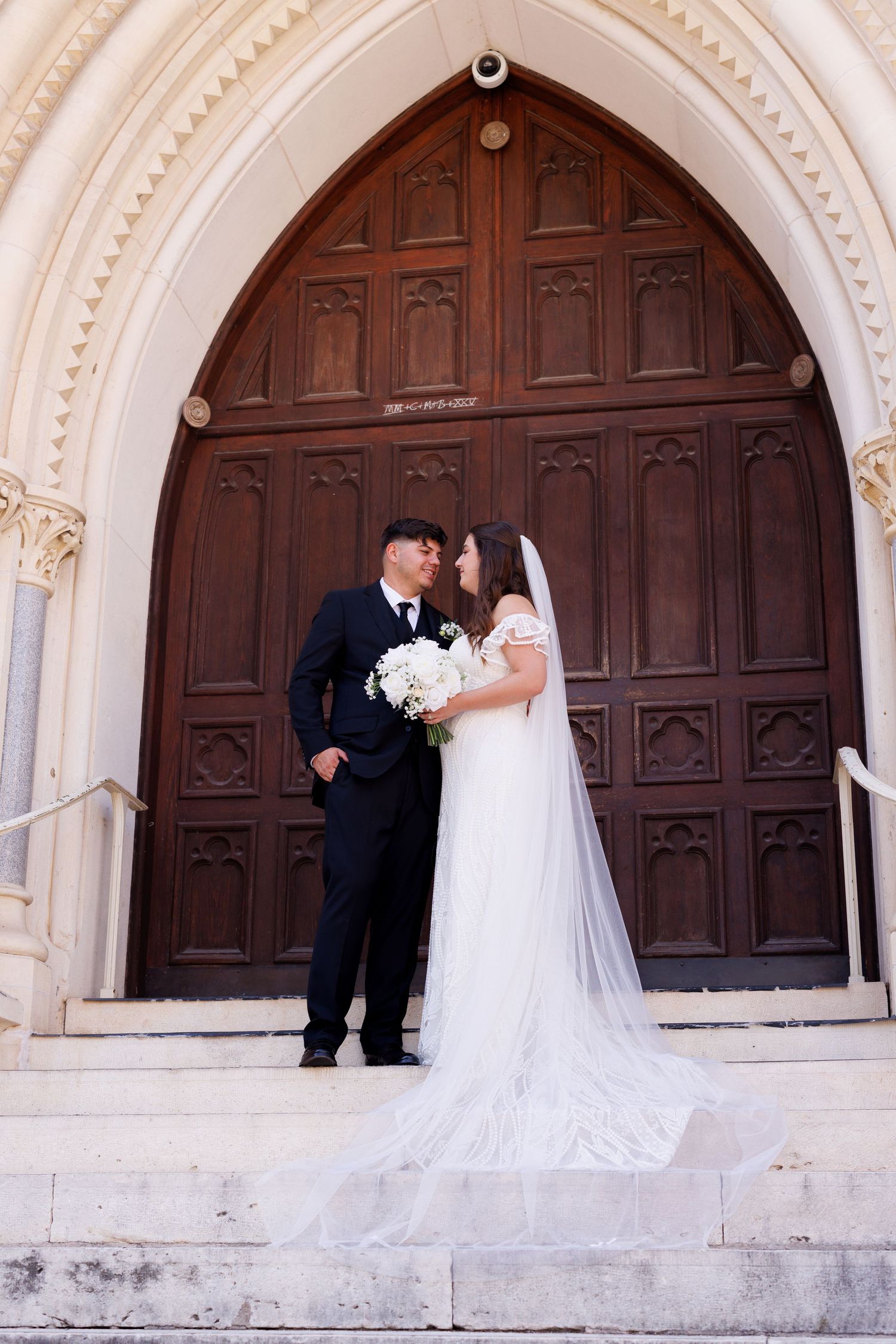 A newlywed couple stands together on church steps before a grand wooden arched doorway.