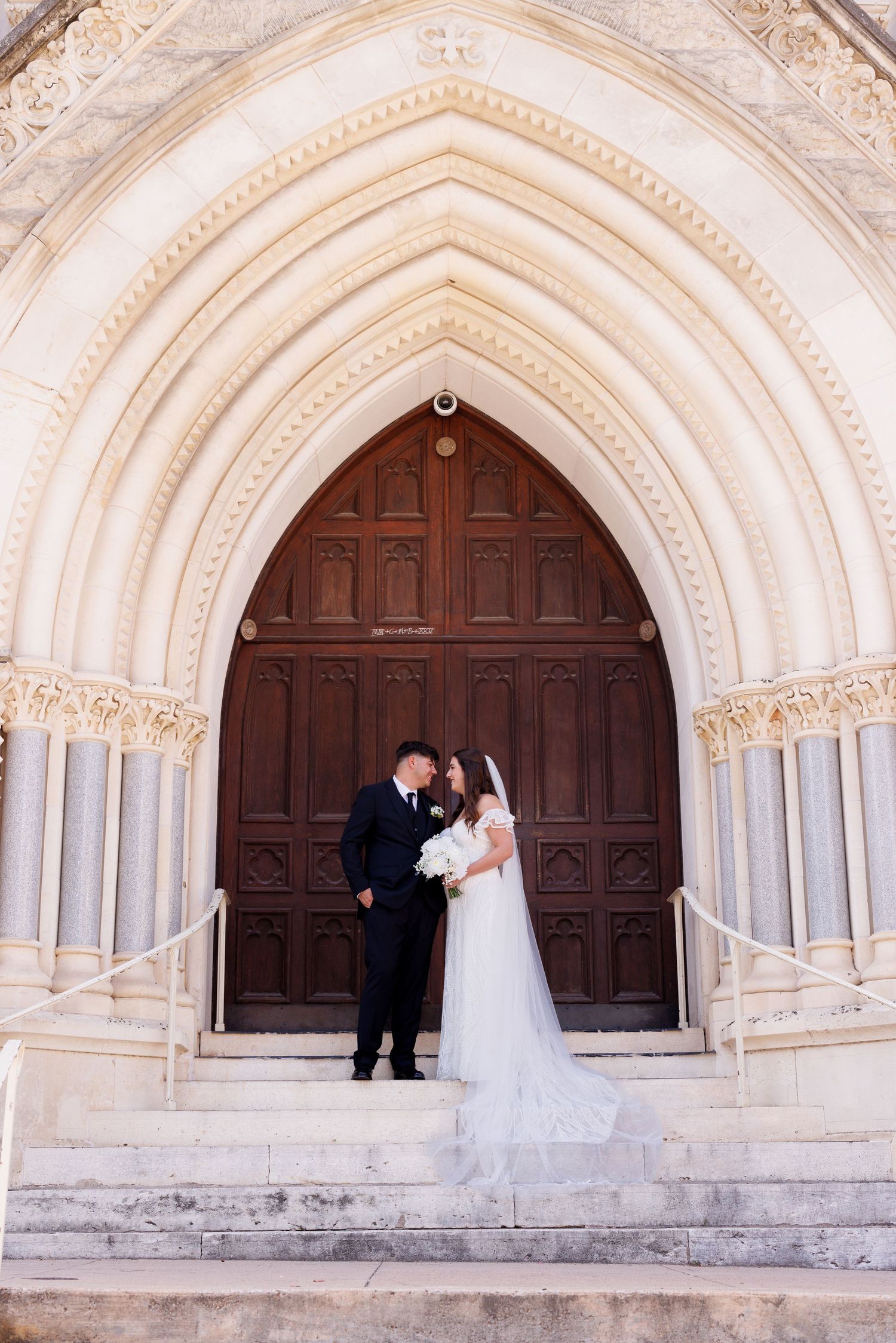 A wedding couple embraces in front of an ornate gothic church entrance with detailed stonework.