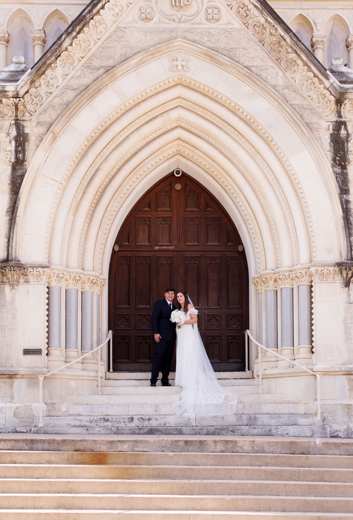 A wedding couple poses beneath an ornate gothic archway of a historic church entrance.