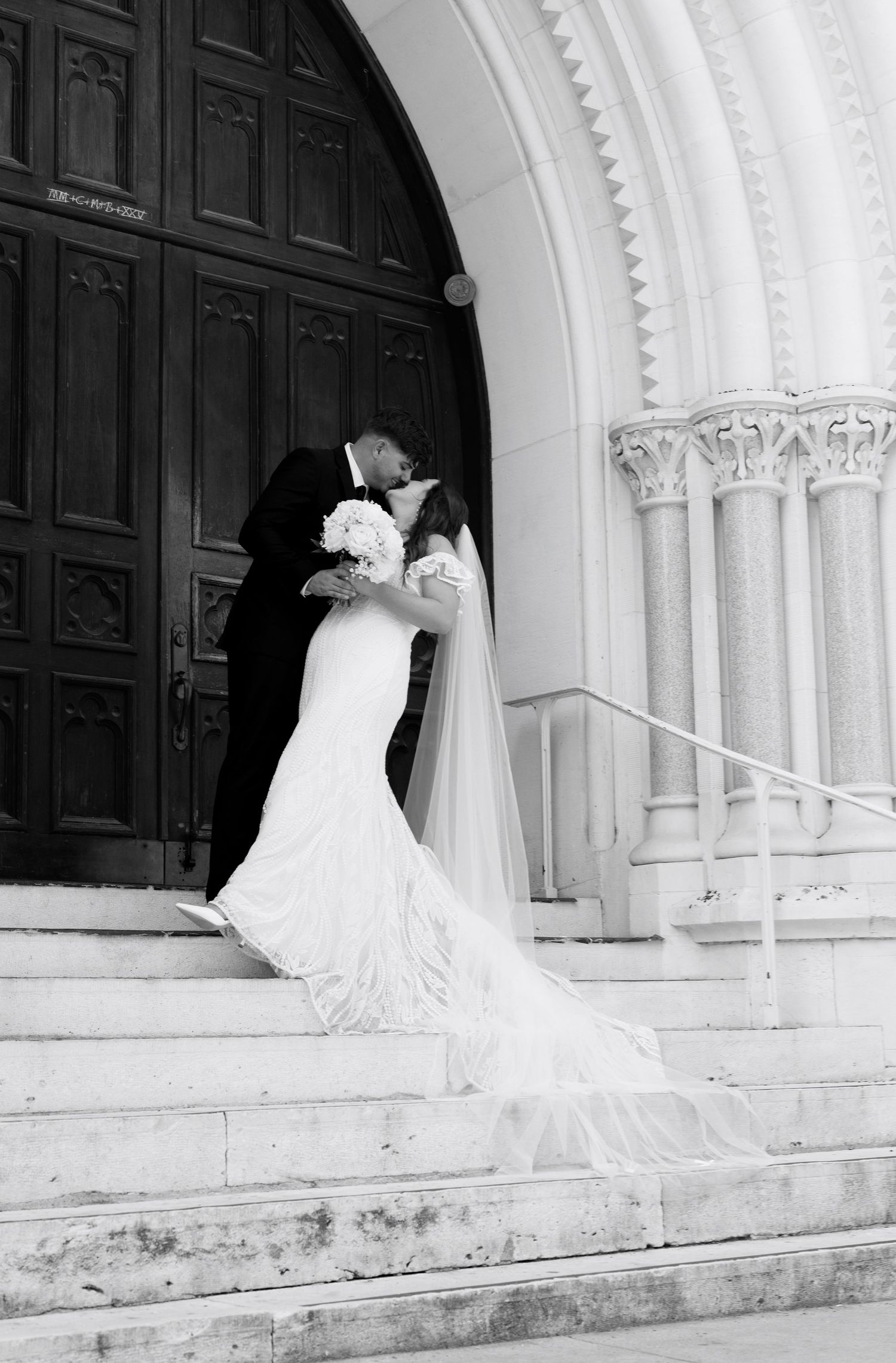 A black and white photo captures a romantic moment between newlyweds on church steps.