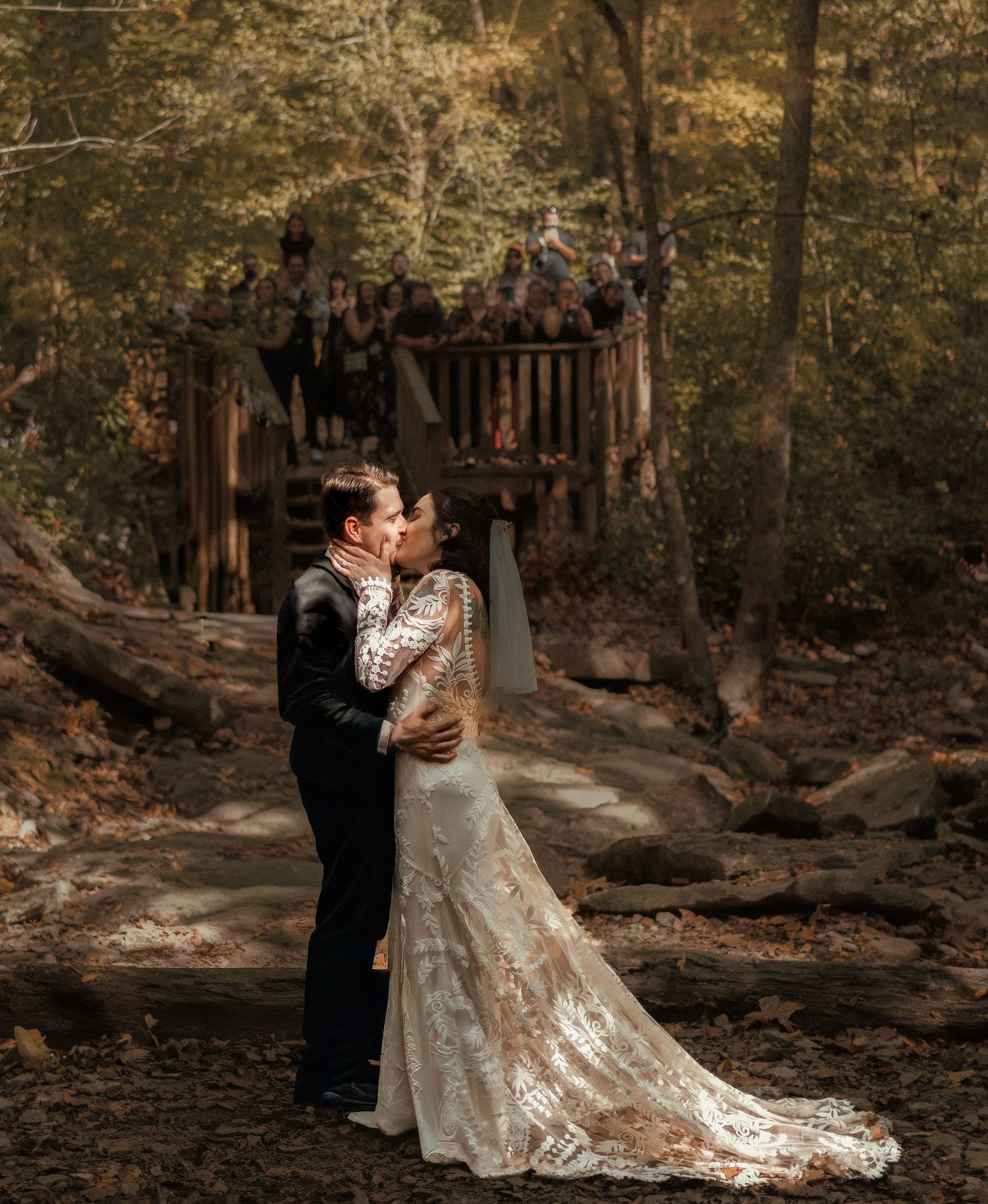 A couple shares a romantic kiss in a forest wedding setting, bride in lace gown with train, guests watching from wooden deck.