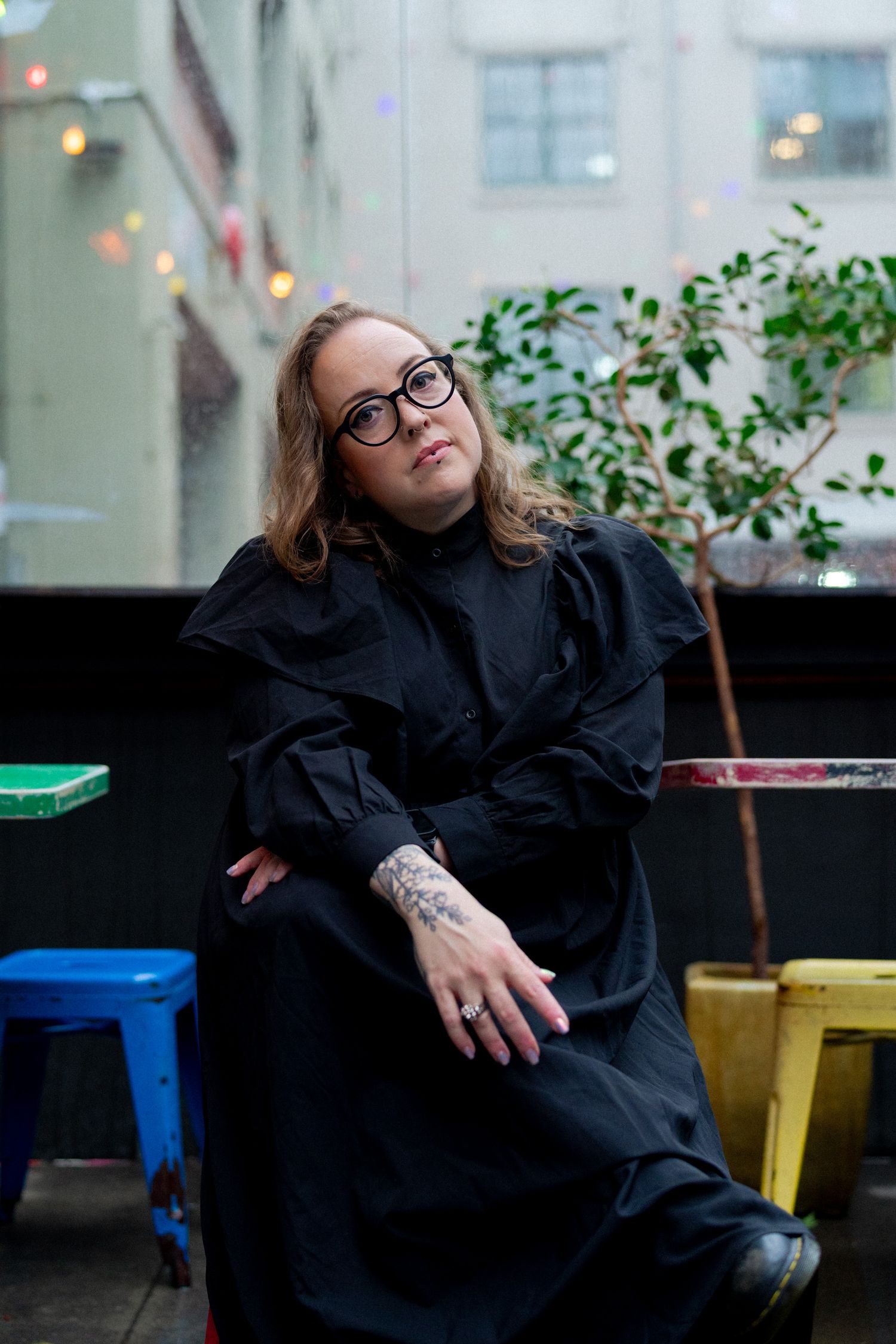 Emma Barnes sits in an outdoor seated area with colourful chairs and festoon lights. They wear a black dress and you can see a tattoo hand.