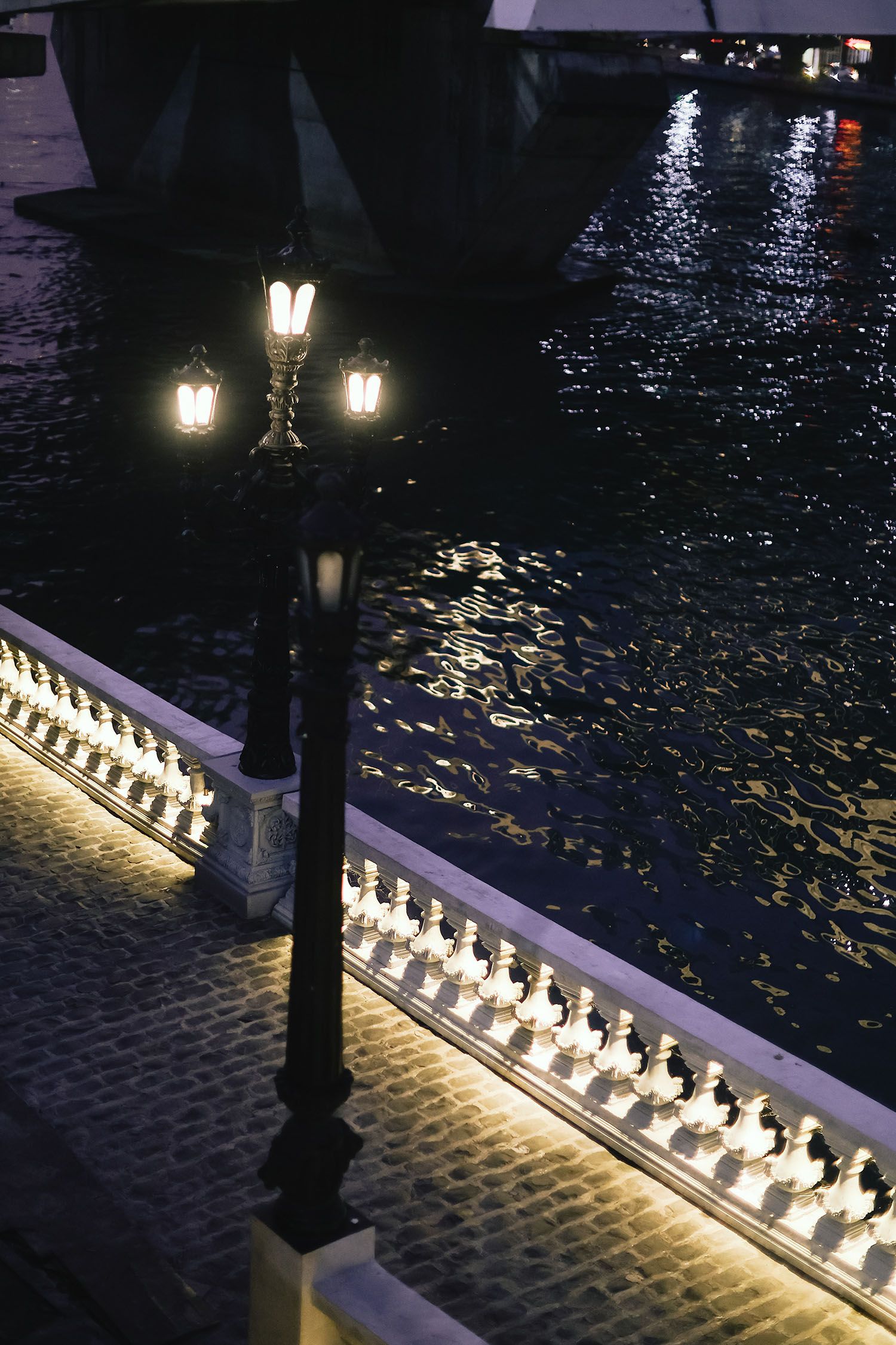 Illuminated white candles line a railing at night, creating a glowing path beside a dark cobblestone walkway.