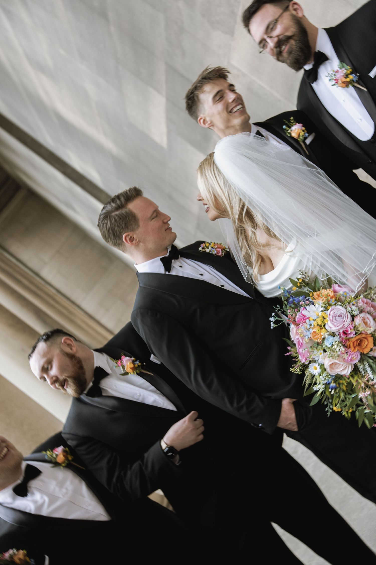 Wedding party in black tuxedos standing together with bride holding colorful bouquet and veil.