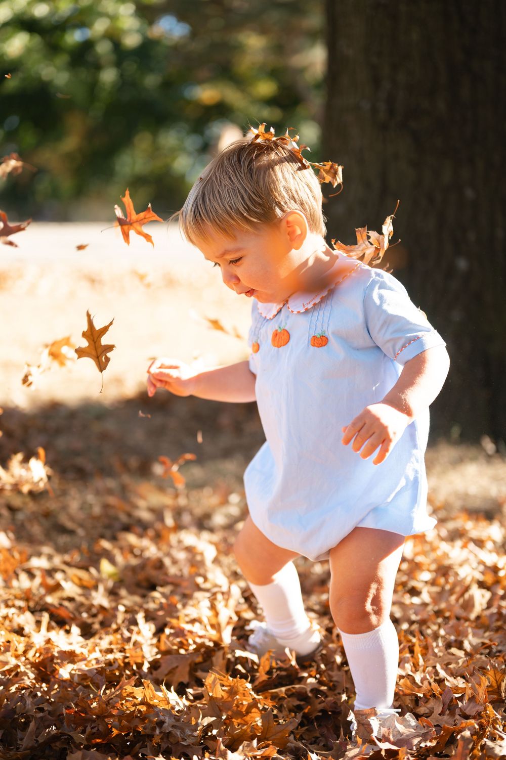 A small child in white clothing plays joyfully in a pile of fallen autumn leaves on a sunny day.
