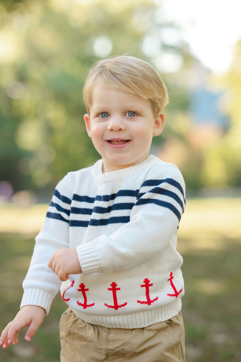 A white knit sweater with red anchor pattern detail is shown in an outdoor setting with natural bokeh background.