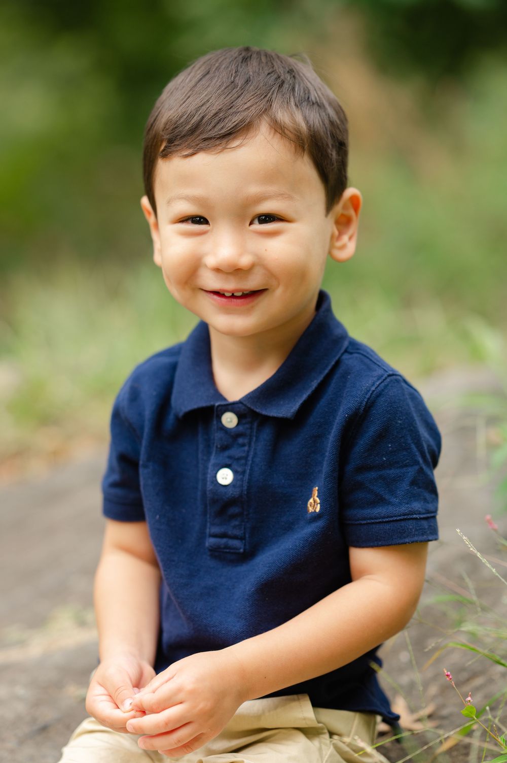 Young child wearing a navy blue polo shirt and khaki pants sits outdoors smiling in a natural setting.