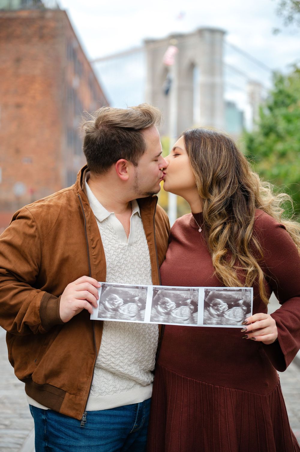 A couple shares a kiss while holding ultrasound photos in a series of romantic outdoor announcement pictures.