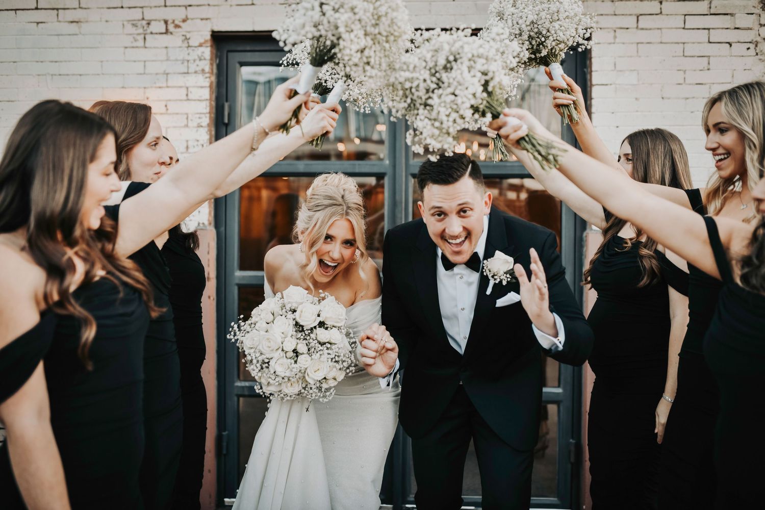 ride and groom laughing as their bridal party lifts bouquets high, creating a colorful floral tunnel at sunset.